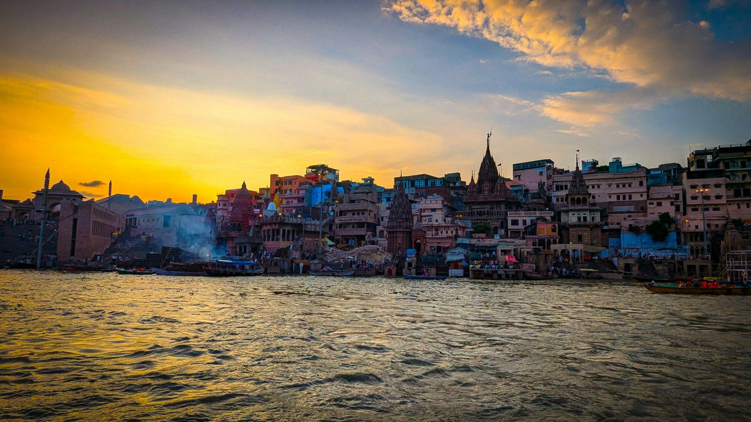 The ghats of Varanasi at sunset, seen from a boat on the river, with temple spires and brightly painted houses along the bank