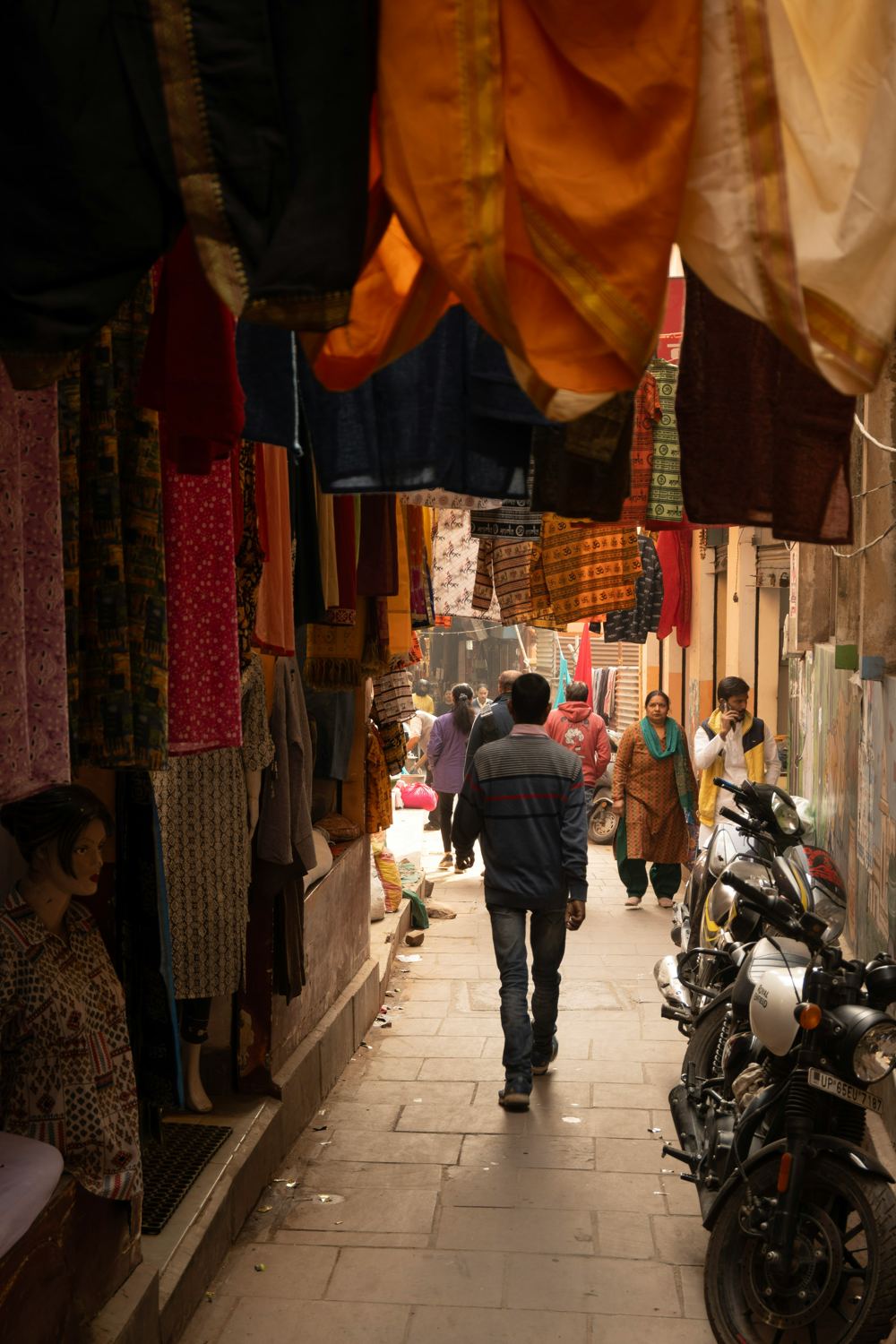 A narrow lane in the old city lined with shops and hanging cloth, with people walking toward the light at the far end